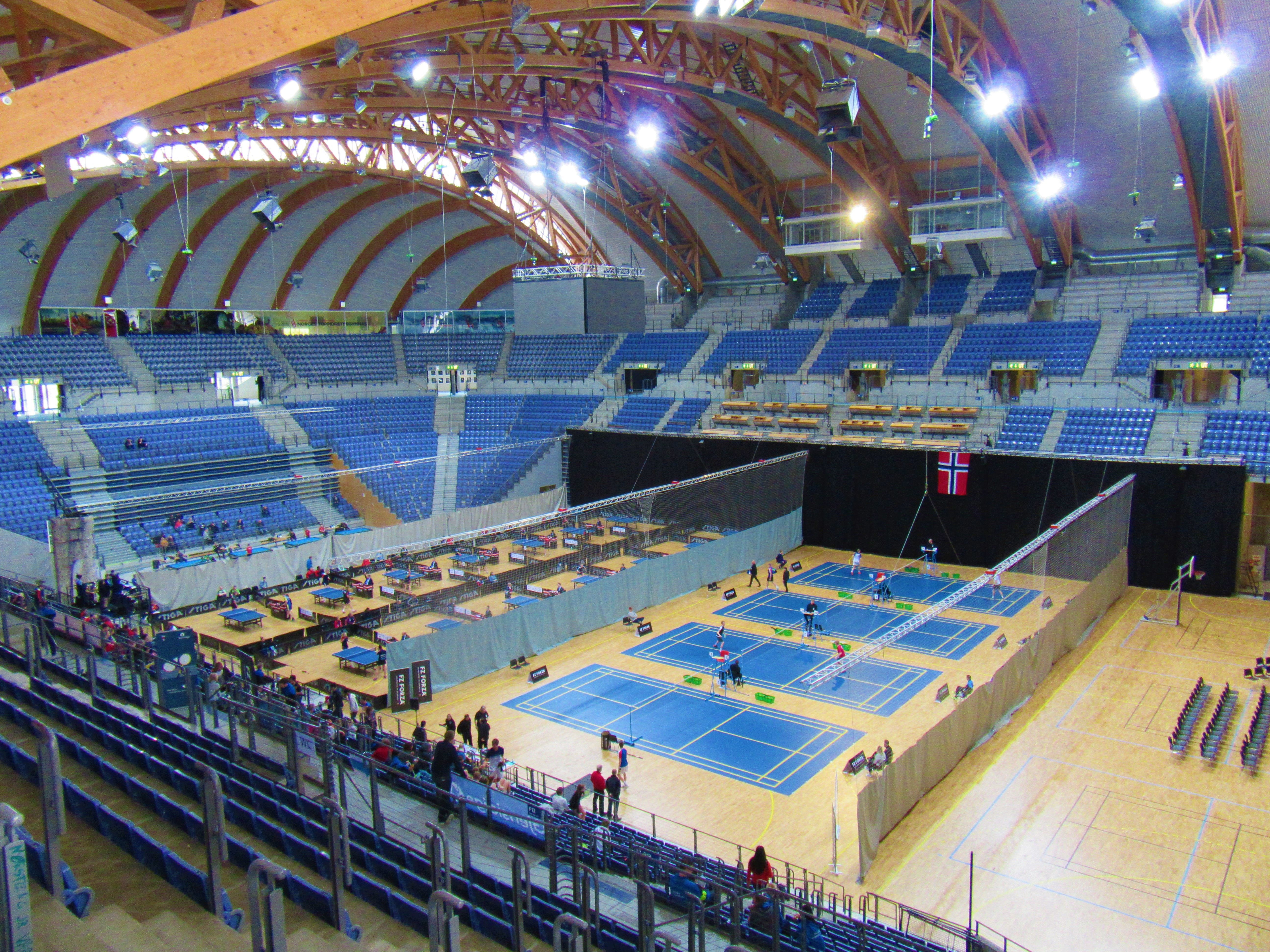 Stor innendørs arena med tribuner og buet tretak, der flere blå baner er satt opp med nett og spillere..