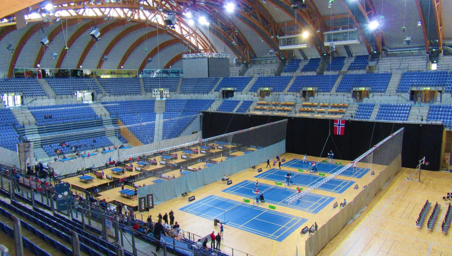 Stor innendørs arena med tribuner og buet tretak, der flere blå baner er satt opp med nett og spillere..
