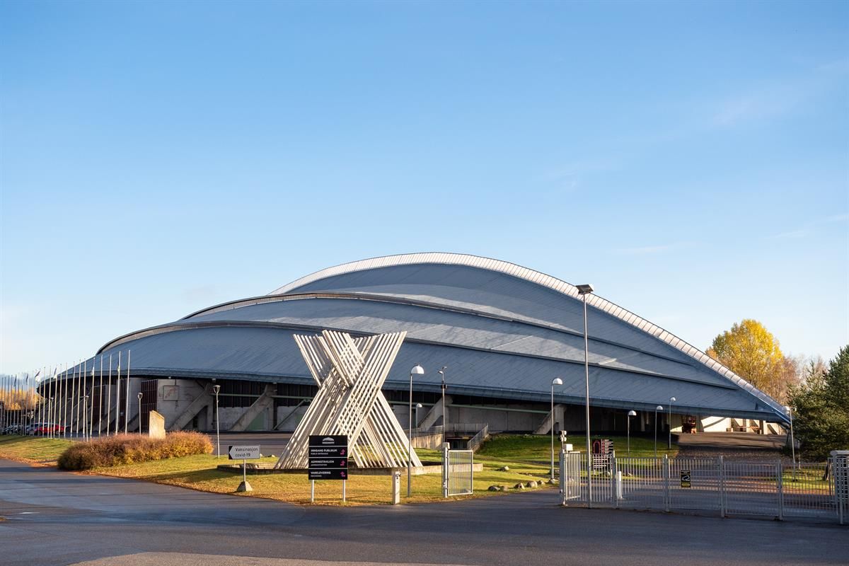 A building with a curved metal roof and a large X‑shaped sculpture in front of the entrance. Sunlight casts warm tones on the surroundings.