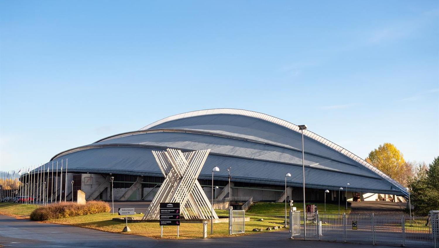 A building with a curved metal roof and a large X‑shaped sculpture in front of the entrance. Sunlight casts warm tones on the surroundings.
