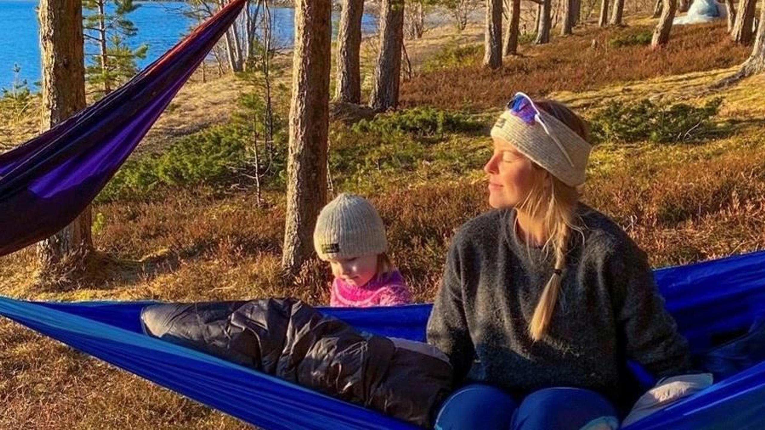 Woman and child sitting in blue hammocks in a sunny pine forest near a lake, wearing warm hats and jackets.