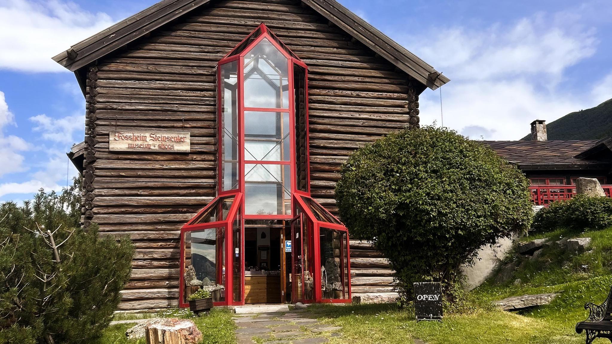 Front view of a wooden log building with a tall red-framed glass entryway, shrubs, and an open sign outside..