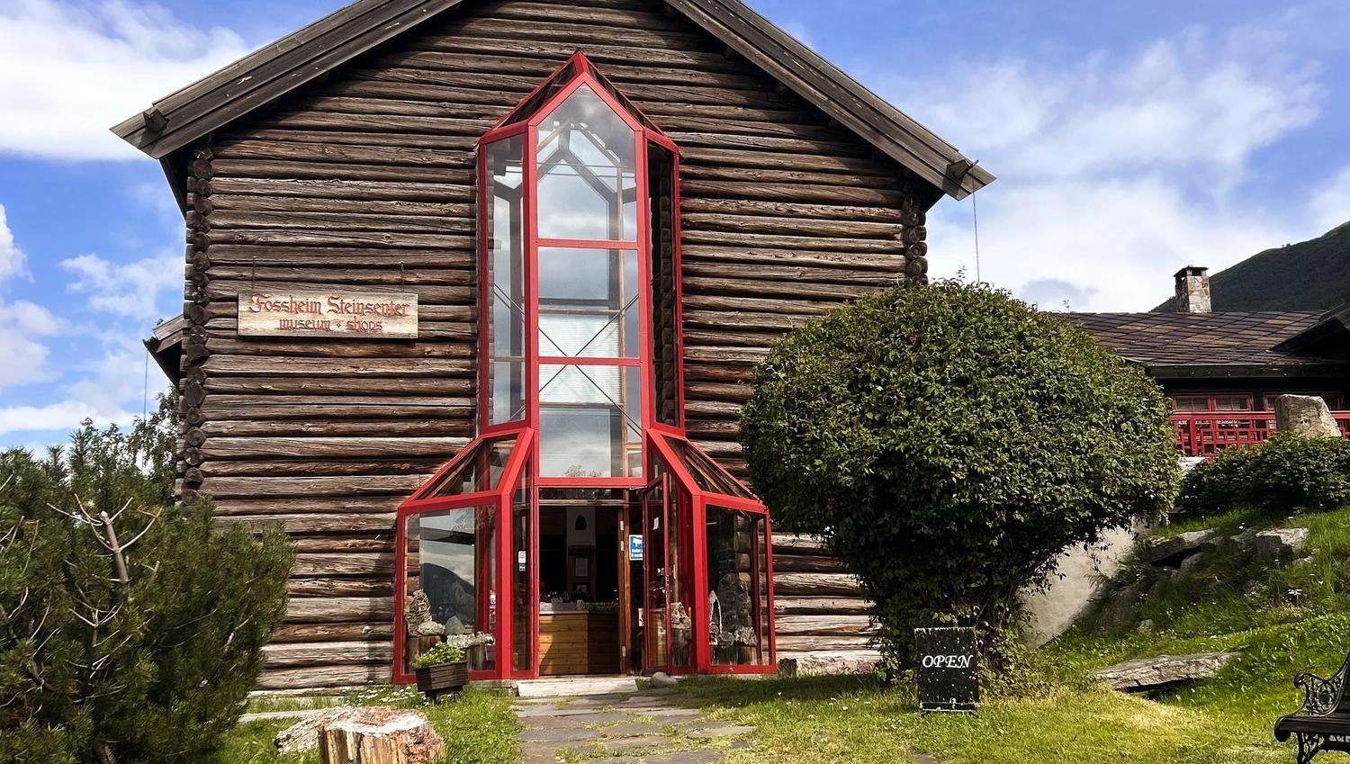 Front view of a wooden log building with a tall red-framed glass entryway, shrubs, and an open sign outside..