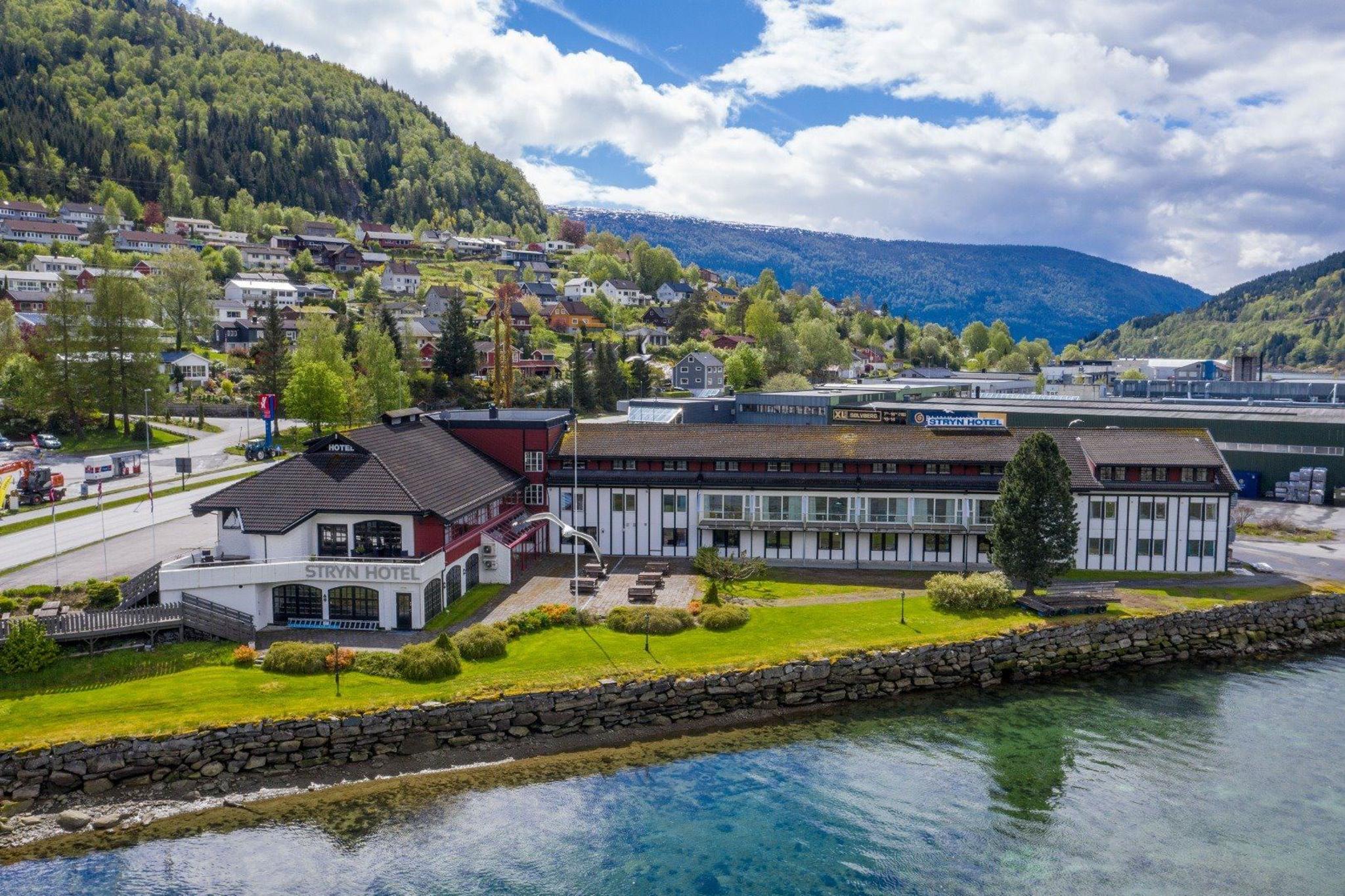 Eit hotellbygg ved fjorden med grøn plen og steinmur, med tettstad og skogkledde fjell under skyer i bakgrunnen..