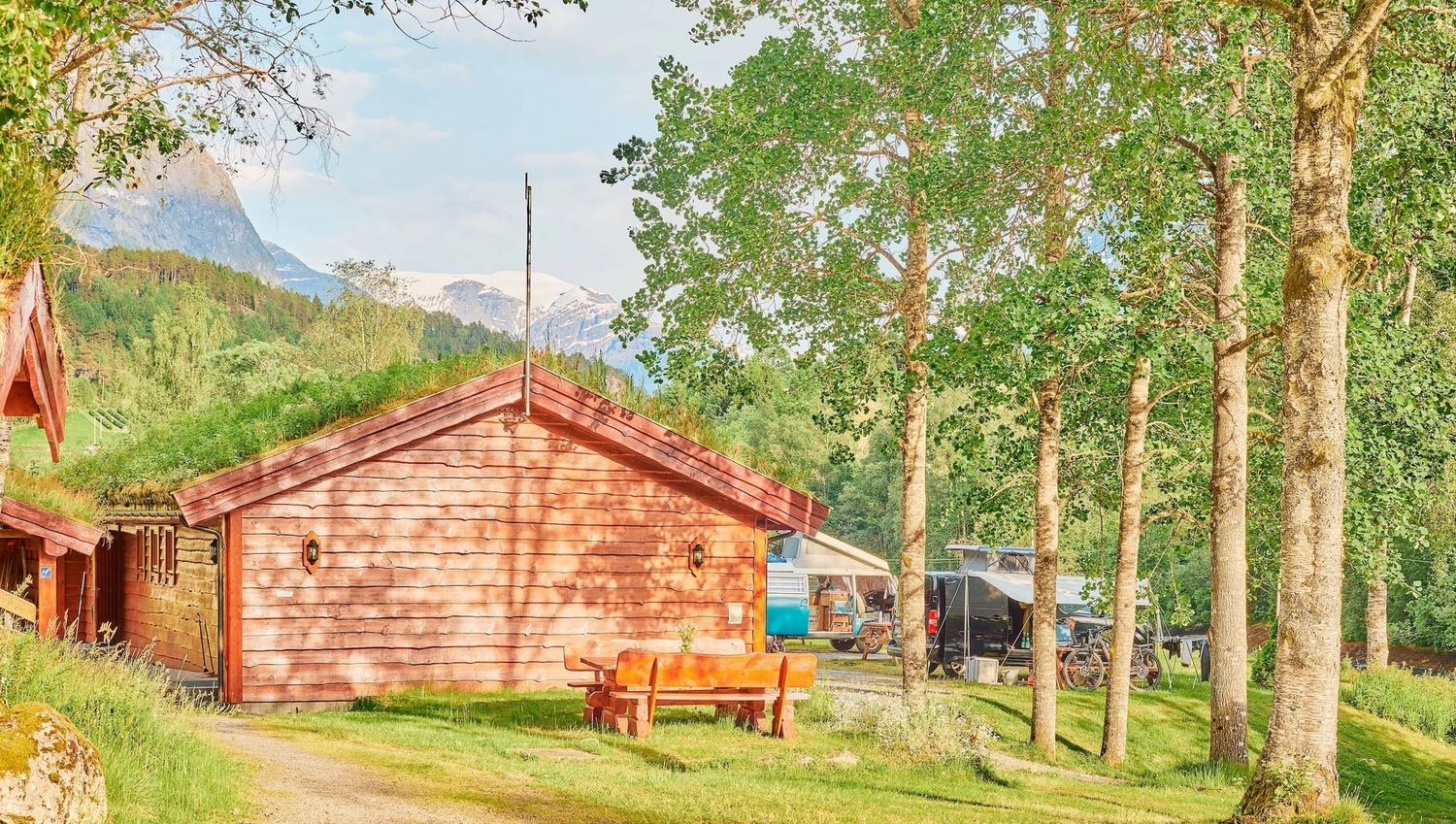 Raud trehytte på grasplen med benk, bjørketrær og campingvogner i bakgrunnen, fjell og blå himmel..