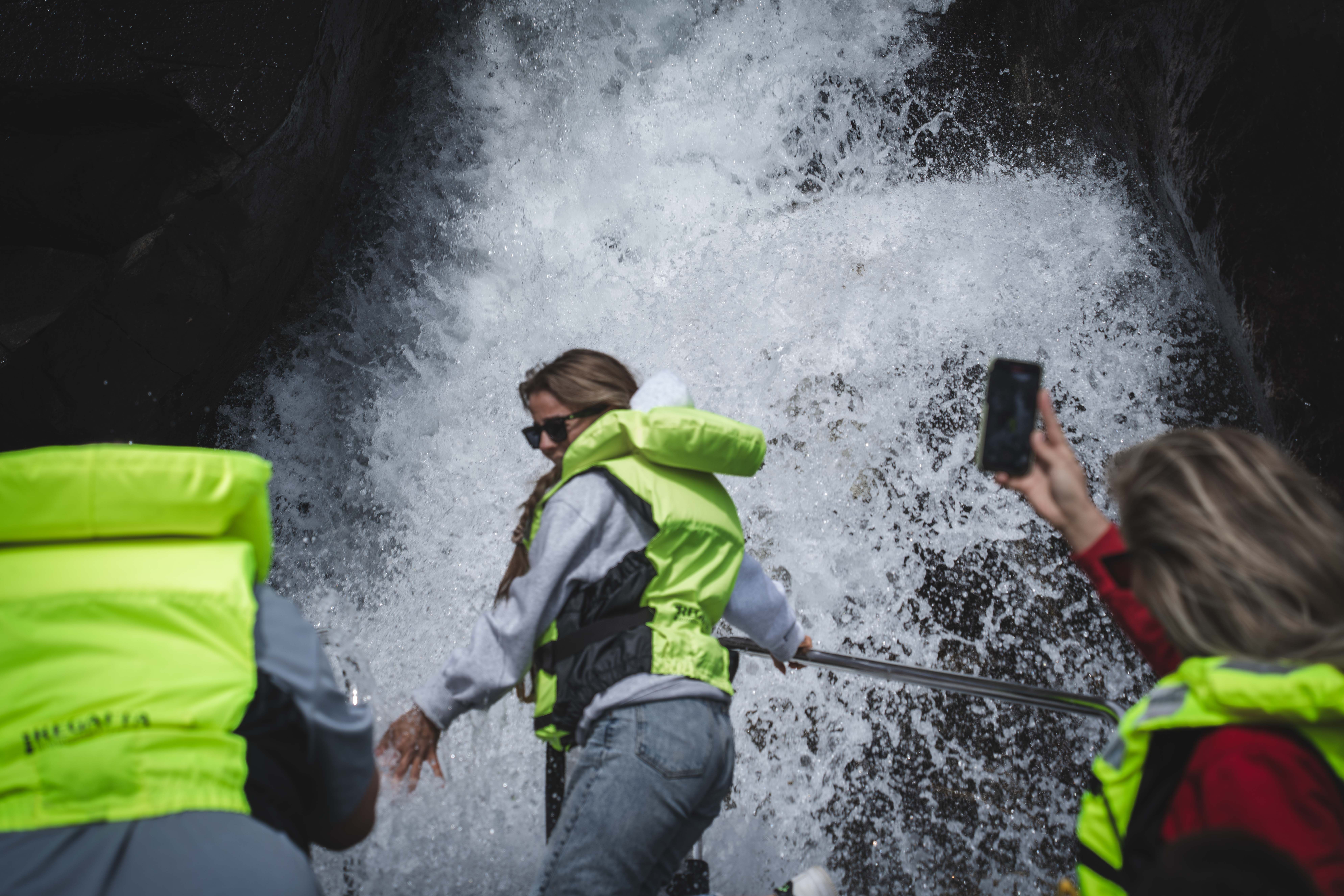 A woman in the front of the boat being "showered" by a waterfall, other passengers are taking photos with their phones.