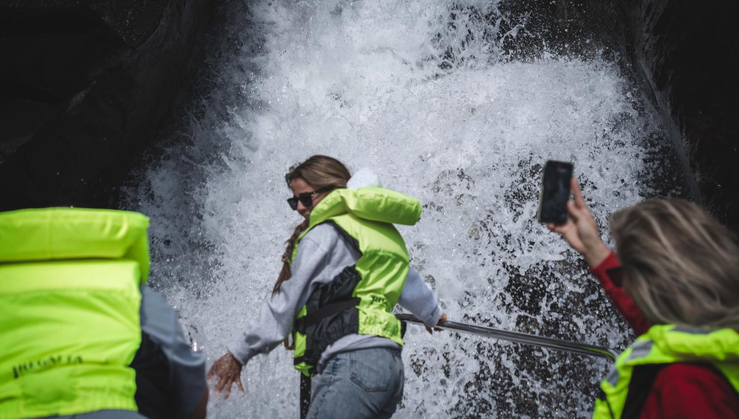 A woman in the front of the boat being "showered" by a waterfall, other passengers are taking photos with their phones.