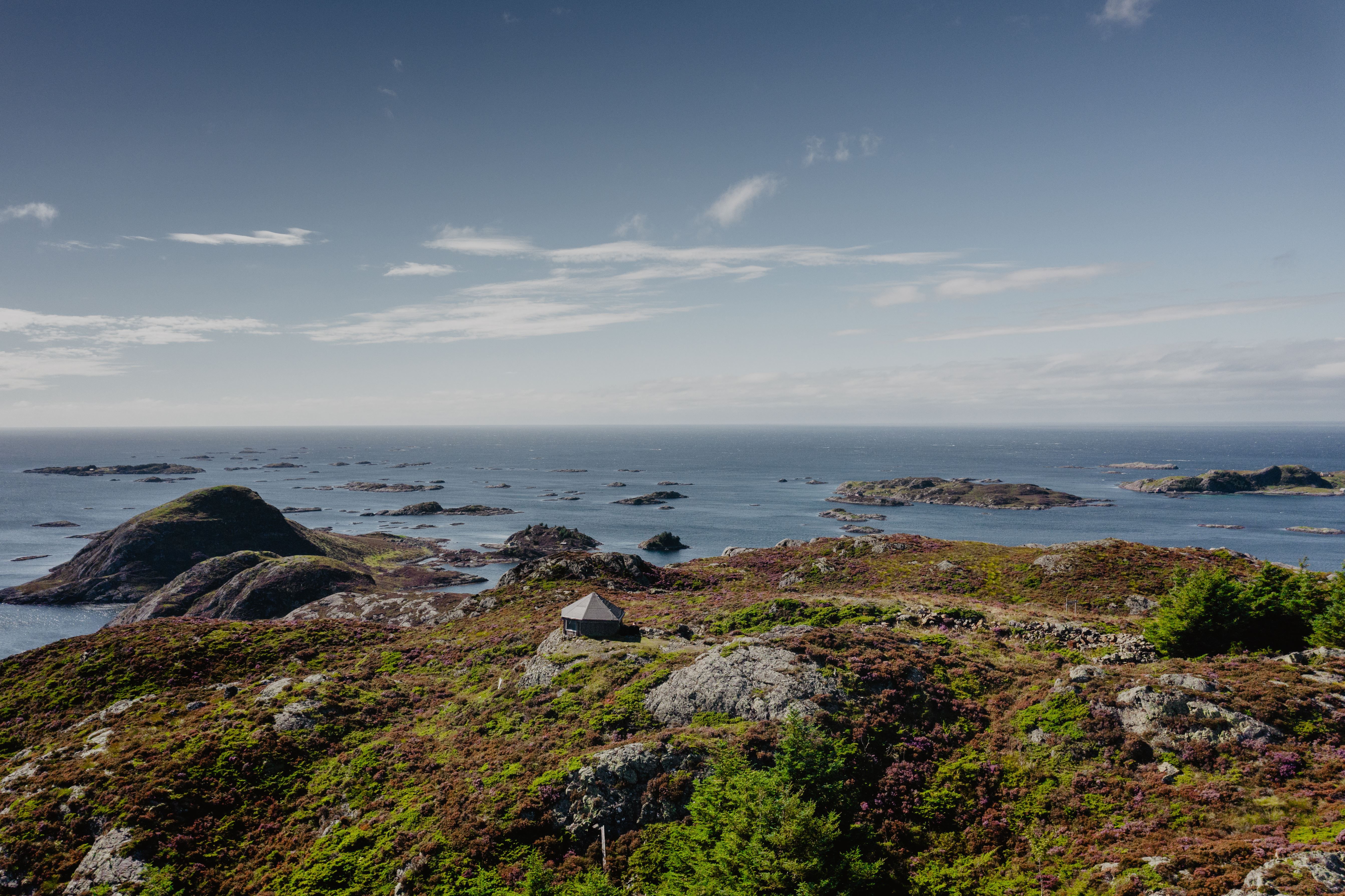 Rocky coastal hillside with a small cabin in the foreground, overlooking a calm sea dotted with small islands.