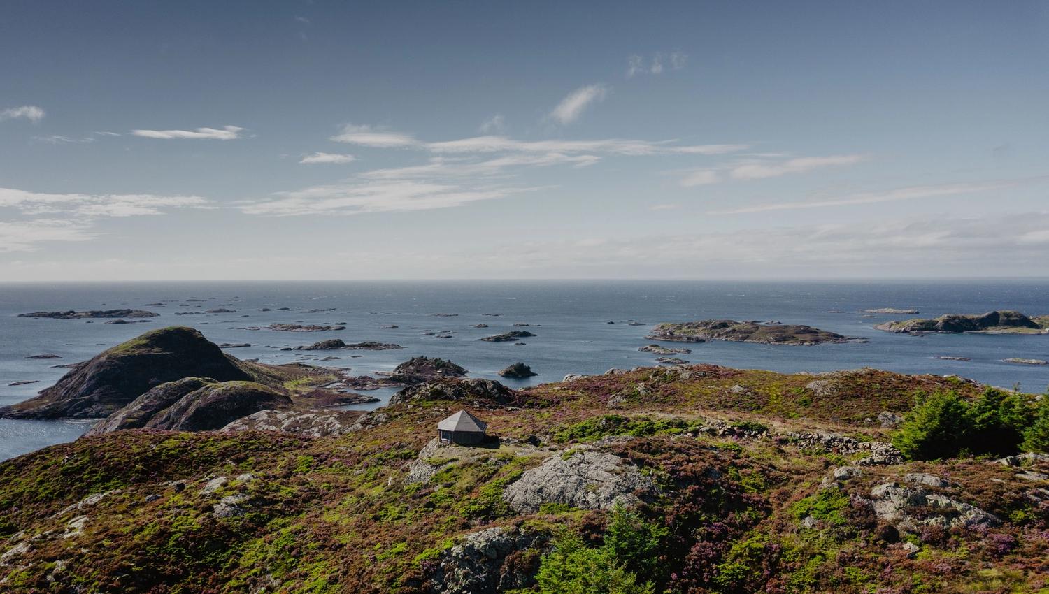 Rocky coastal hillside with a small cabin in the foreground, overlooking a calm sea dotted with small islands.