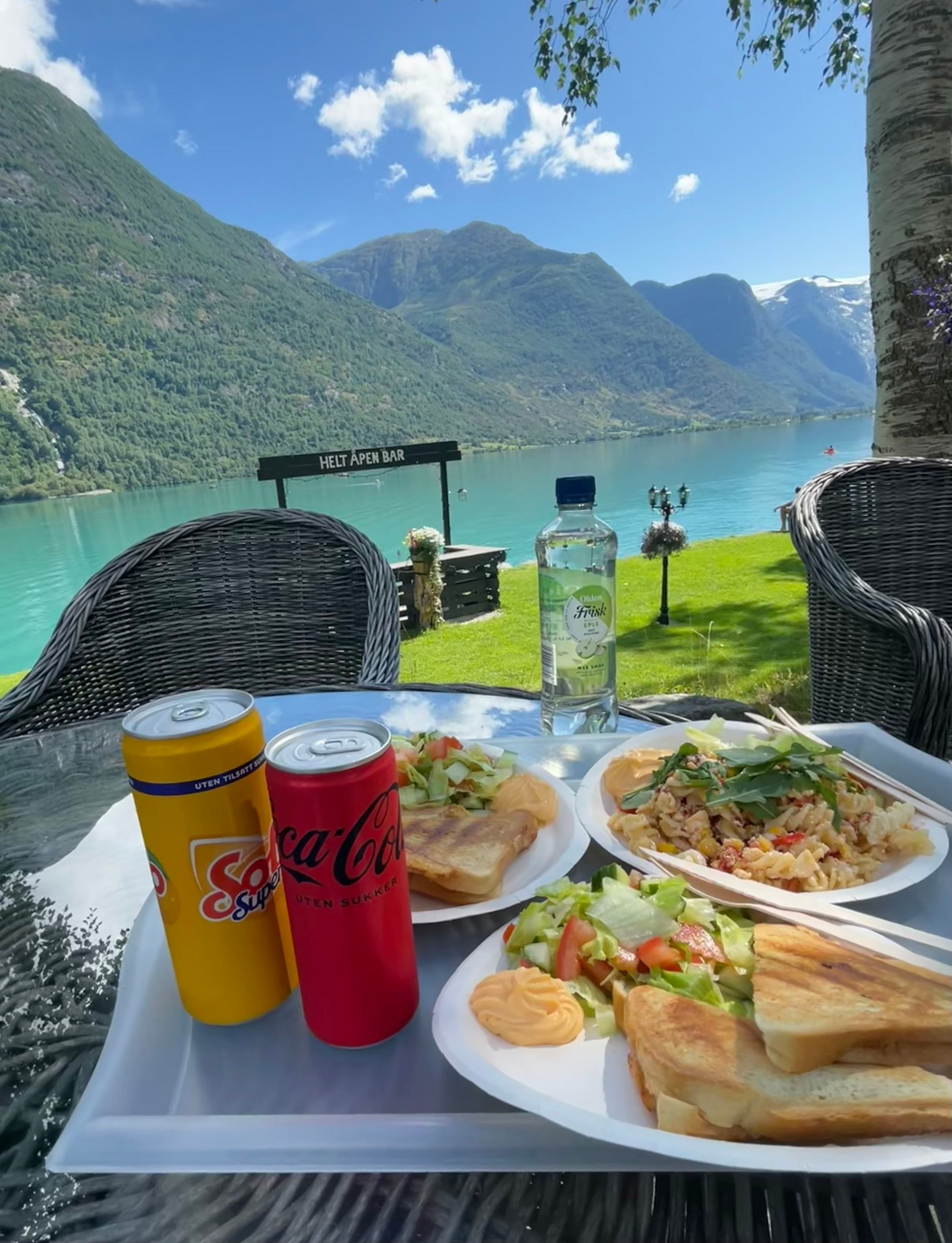 Outdoor table with sandwiches, salads, pasta, soda cans and water bottle, overlooking a turquoise lake and mountains.