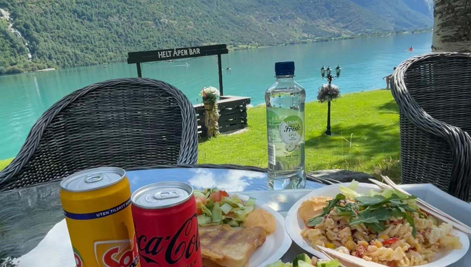Outdoor table with sandwiches, salads, pasta, soda cans and water bottle, overlooking a turquoise lake and mountains.