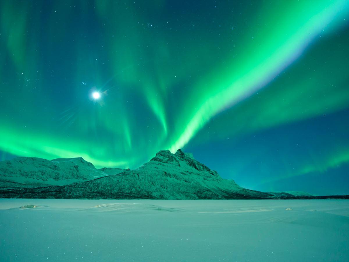 Green Northern Lights dance over snow-capped mountains and a flat, white winter landscape during a starry night.