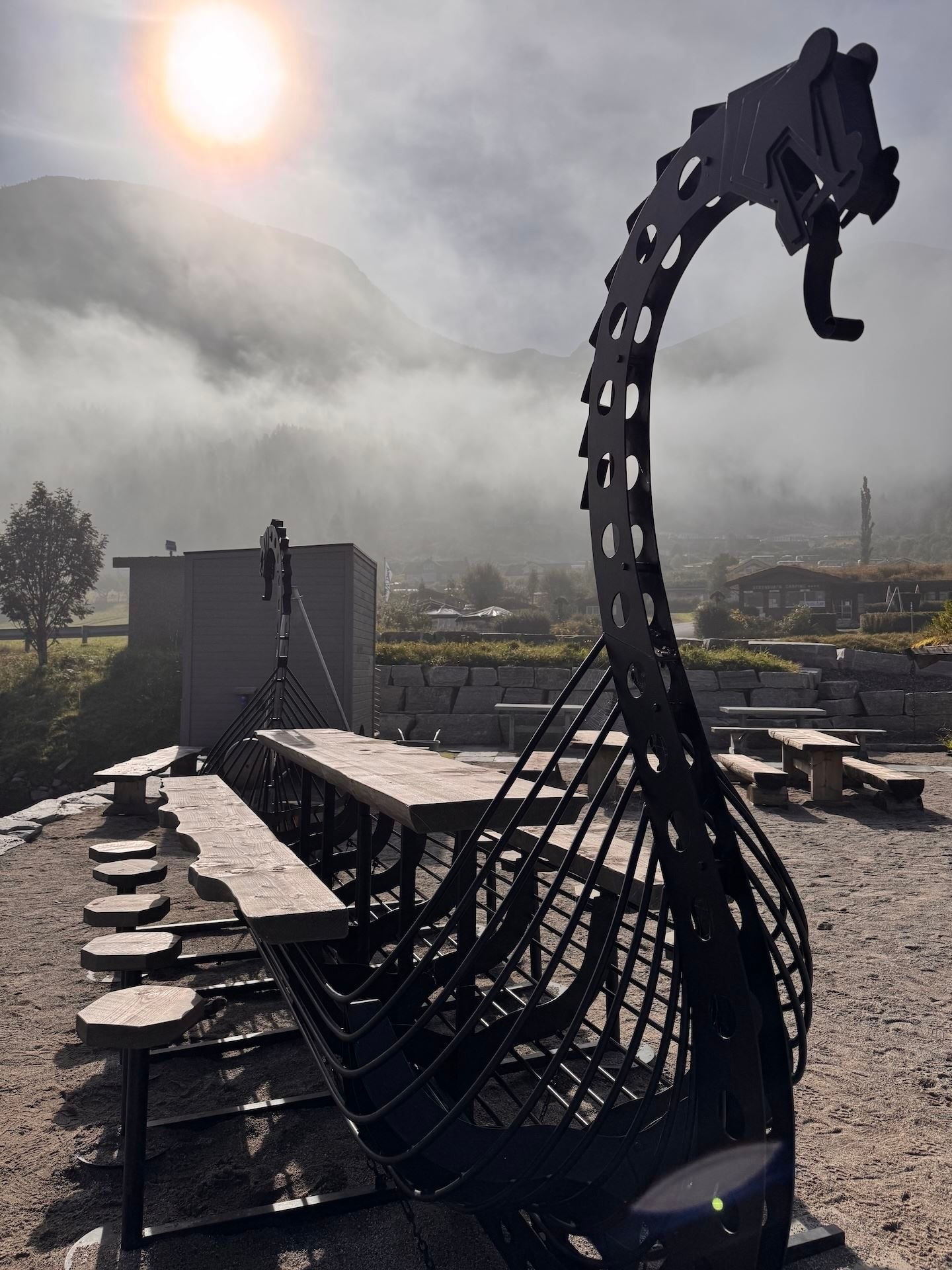 Large black Viking ship-style playground structure beside picnic tables, with misty mountains and sun in sky..