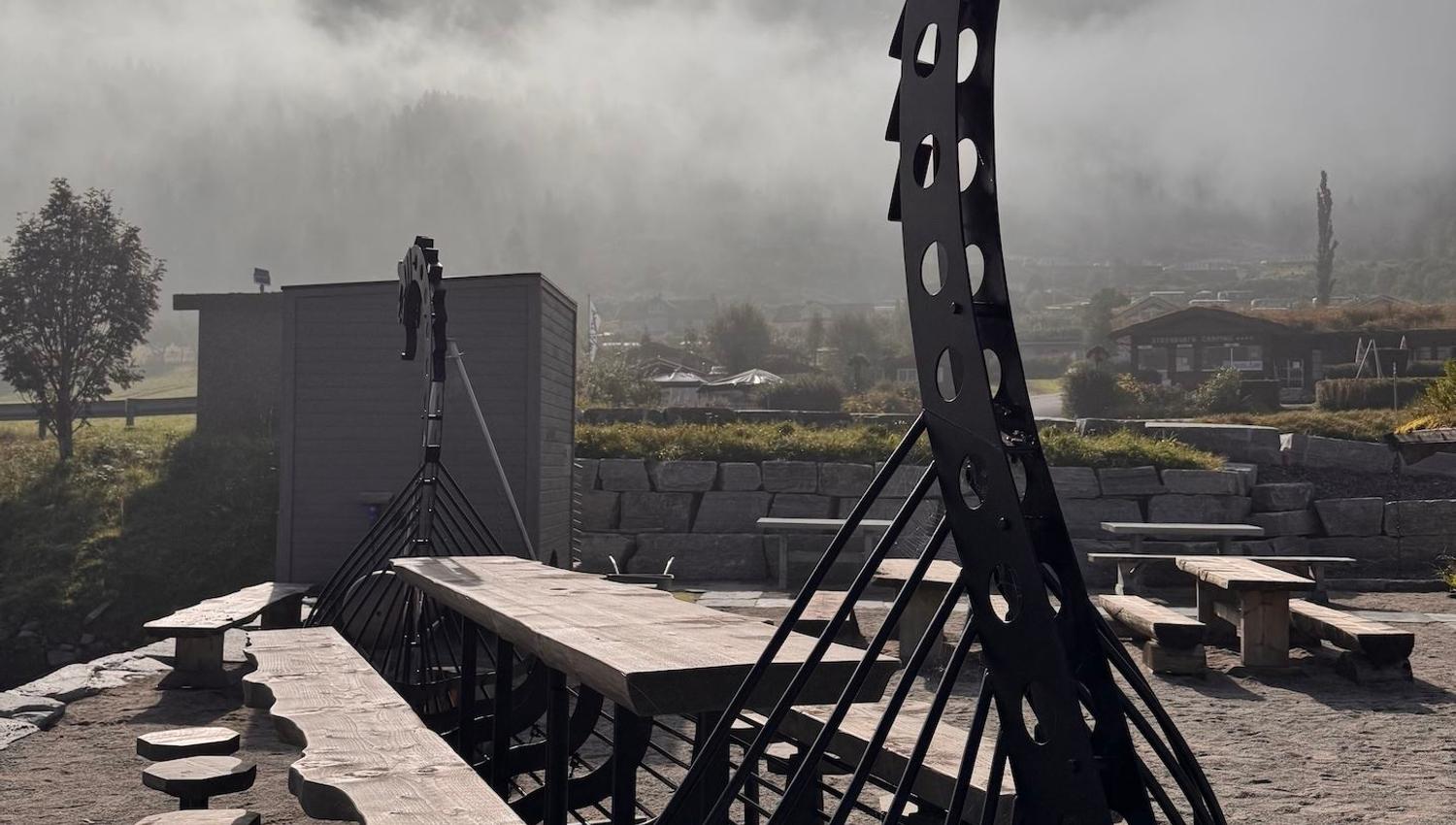 Large black Viking ship-style playground structure beside picnic tables, with misty mountains and sun in sky..