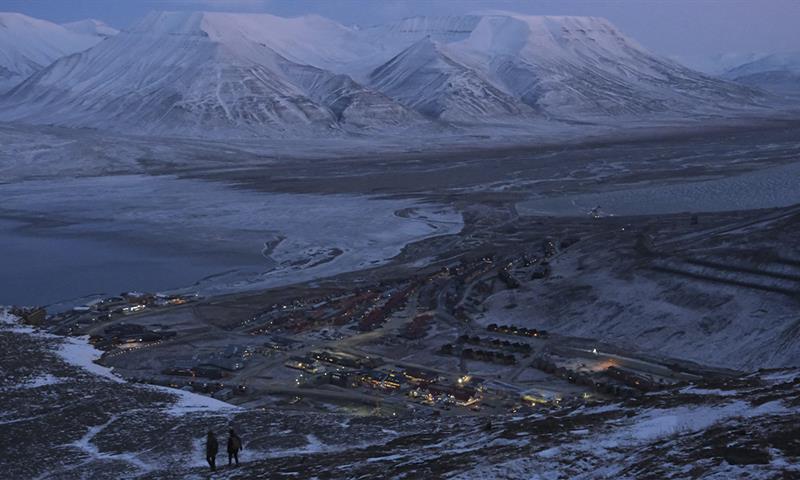 Longyearbyen and nearby snow-covered mountains seen from Platåfjellet with two persons hiking down a hill in the foreground