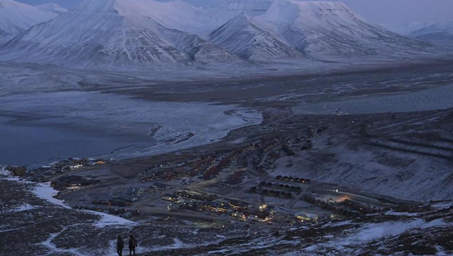 Longyearbyen and nearby snow-covered mountains seen from Platåfjellet with two persons hiking down a hill in the foreground