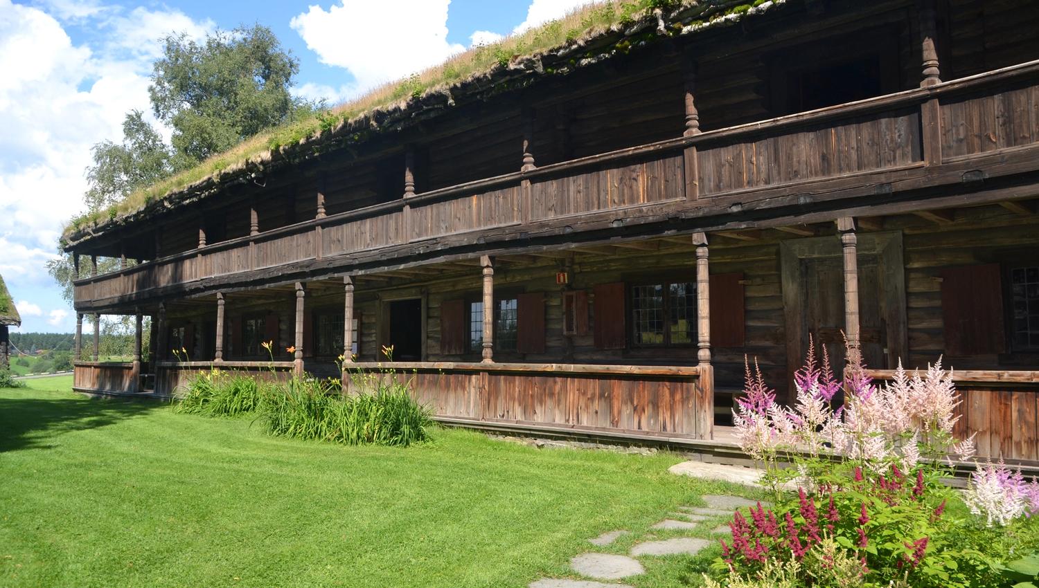 Gamle Hvam svalgang, i brunbrent materialet i sommerdagen med grønt gress og blomstrende stauder. Gamle Hvam