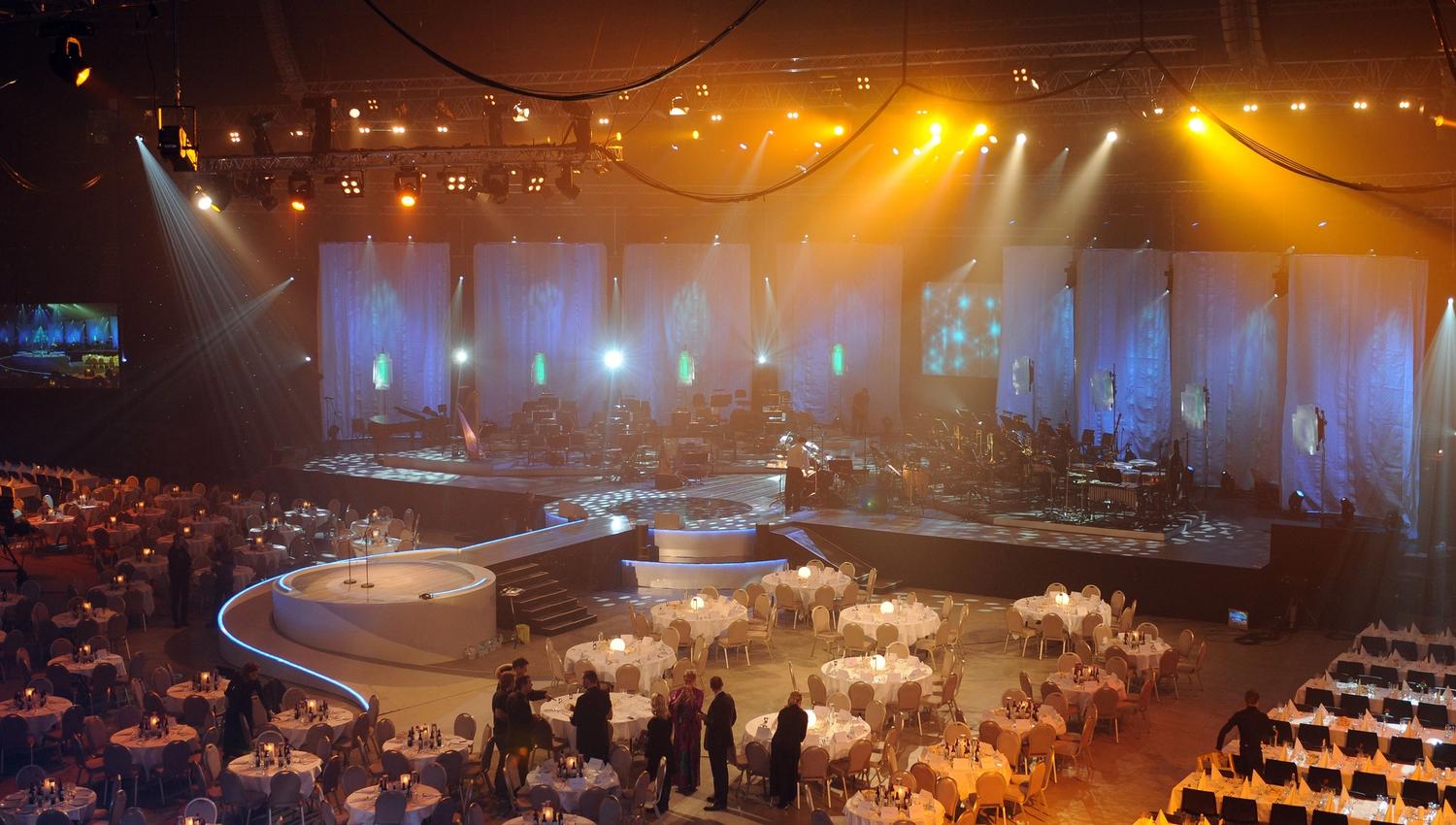 Stor innendørs konsertsal med opplyst scene, instrumenter og runde bord med duker og gjester i salen..