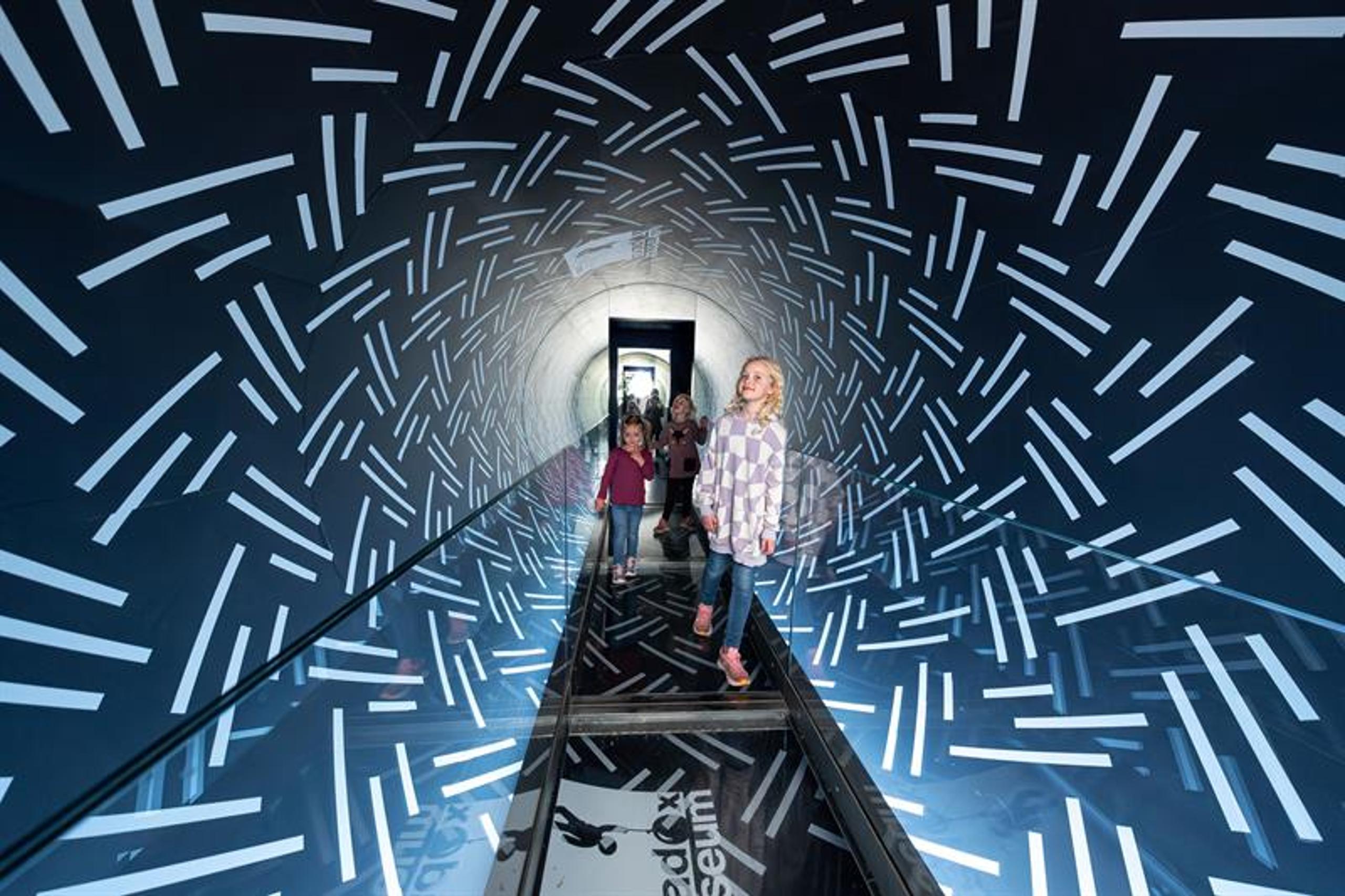 Children walk through a mirrored tunnel with light patterns, creating a futuristic and immersive experience.