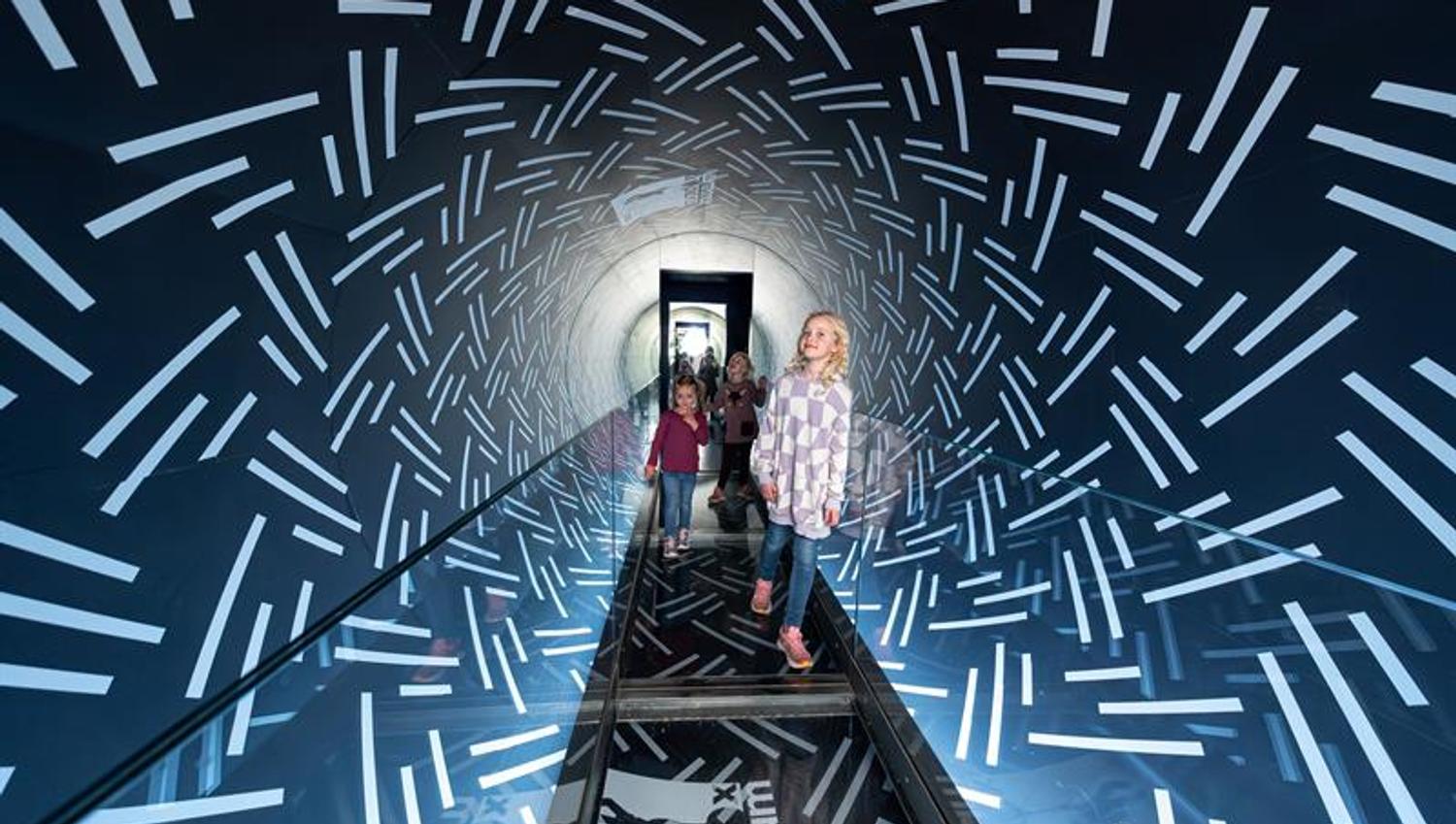 Children walk through a mirrored tunnel with light patterns, creating a futuristic and immersive experience.