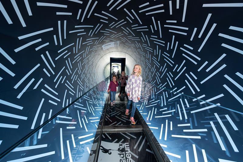 Children walk through a mirrored tunnel with light patterns, creating a futuristic and immersive experience.