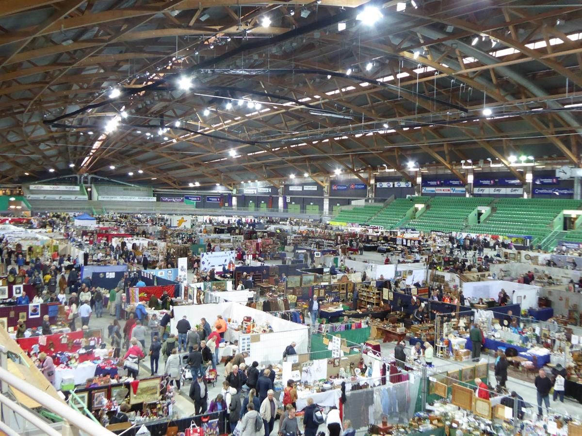 Large indoor exhibition area with many booths, white tents, and visitors. Wooden roof structure and bright ceiling lights fill the hall.