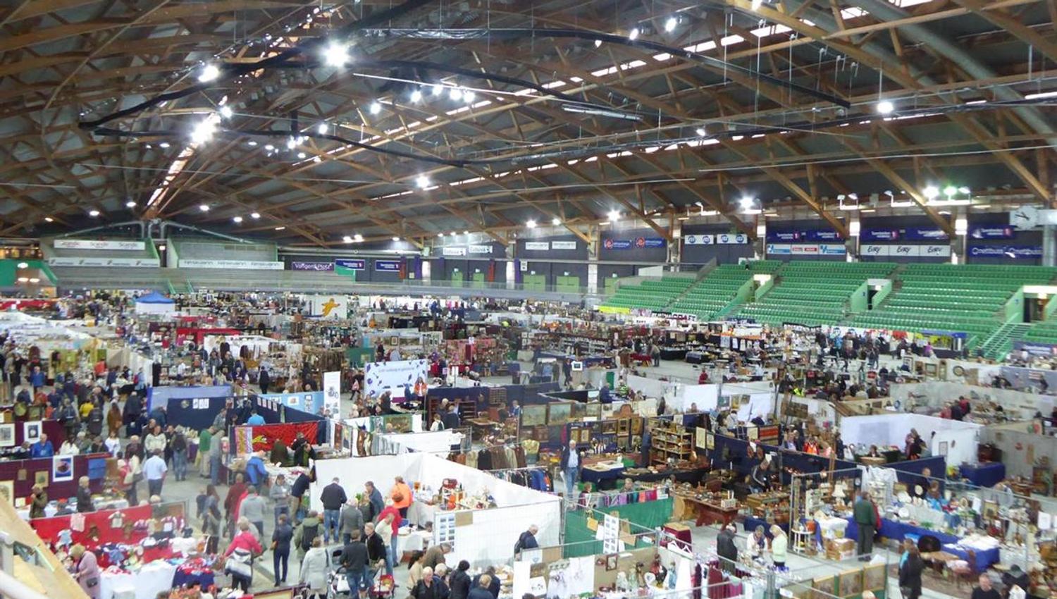 Large indoor exhibition area with many booths, white tents, and visitors. Wooden roof structure and bright ceiling lights fill the hall.