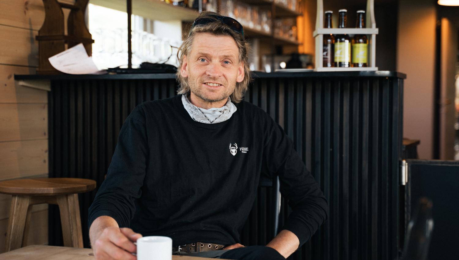 The owner at Halne mountain lodge, sitting at a table holding a cup of coffee.