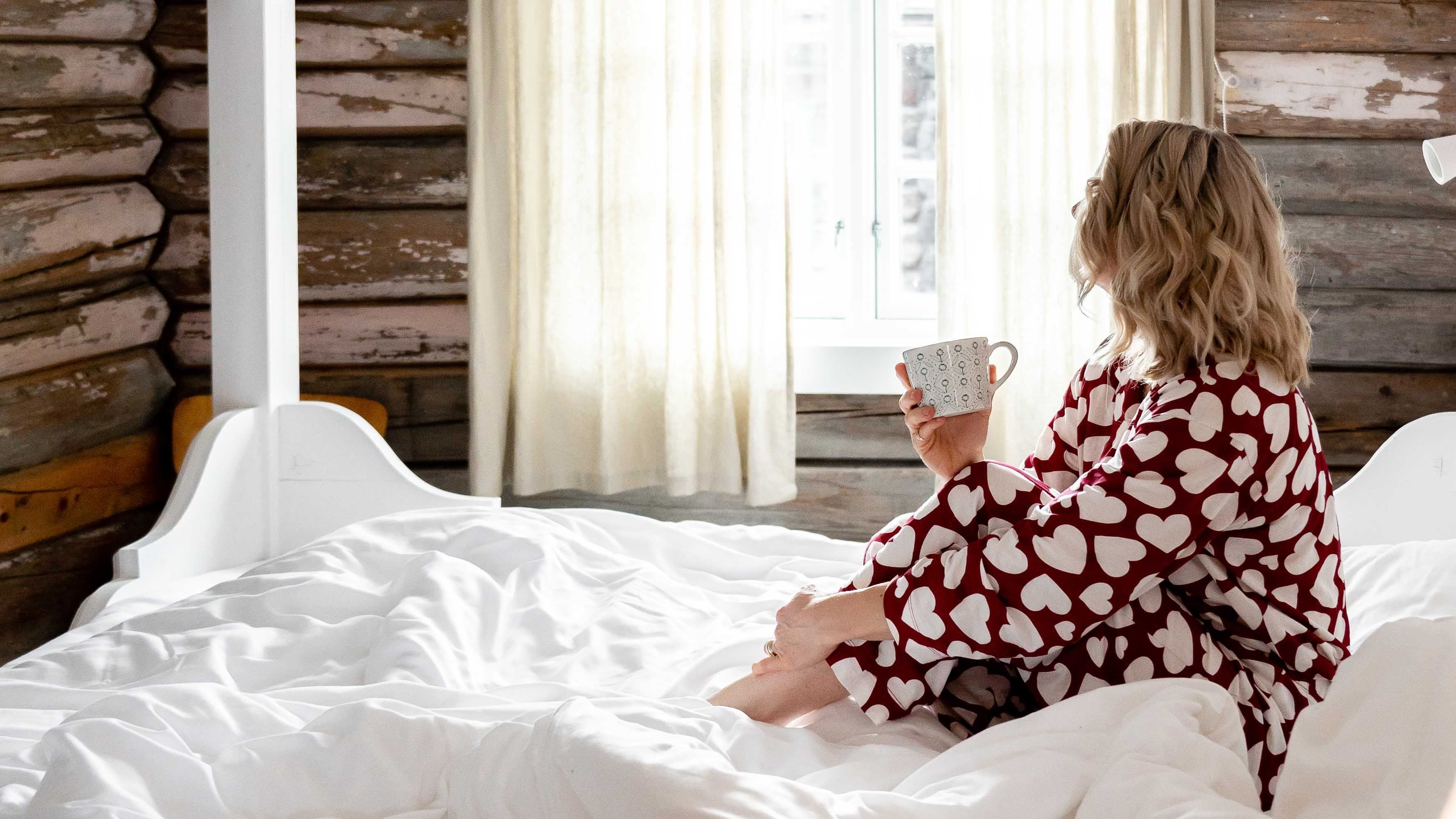Vrouw in hartpatroon-pyjama zit op een wit bed met een mok in haar hand, kijkend naar een helder raam in een blokhutkamer...