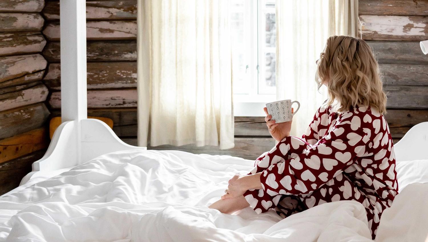 Vrouw in hartpatroon-pyjama zit op een wit bed met een mok in haar hand, kijkend naar een helder raam in een blokhutkamer...