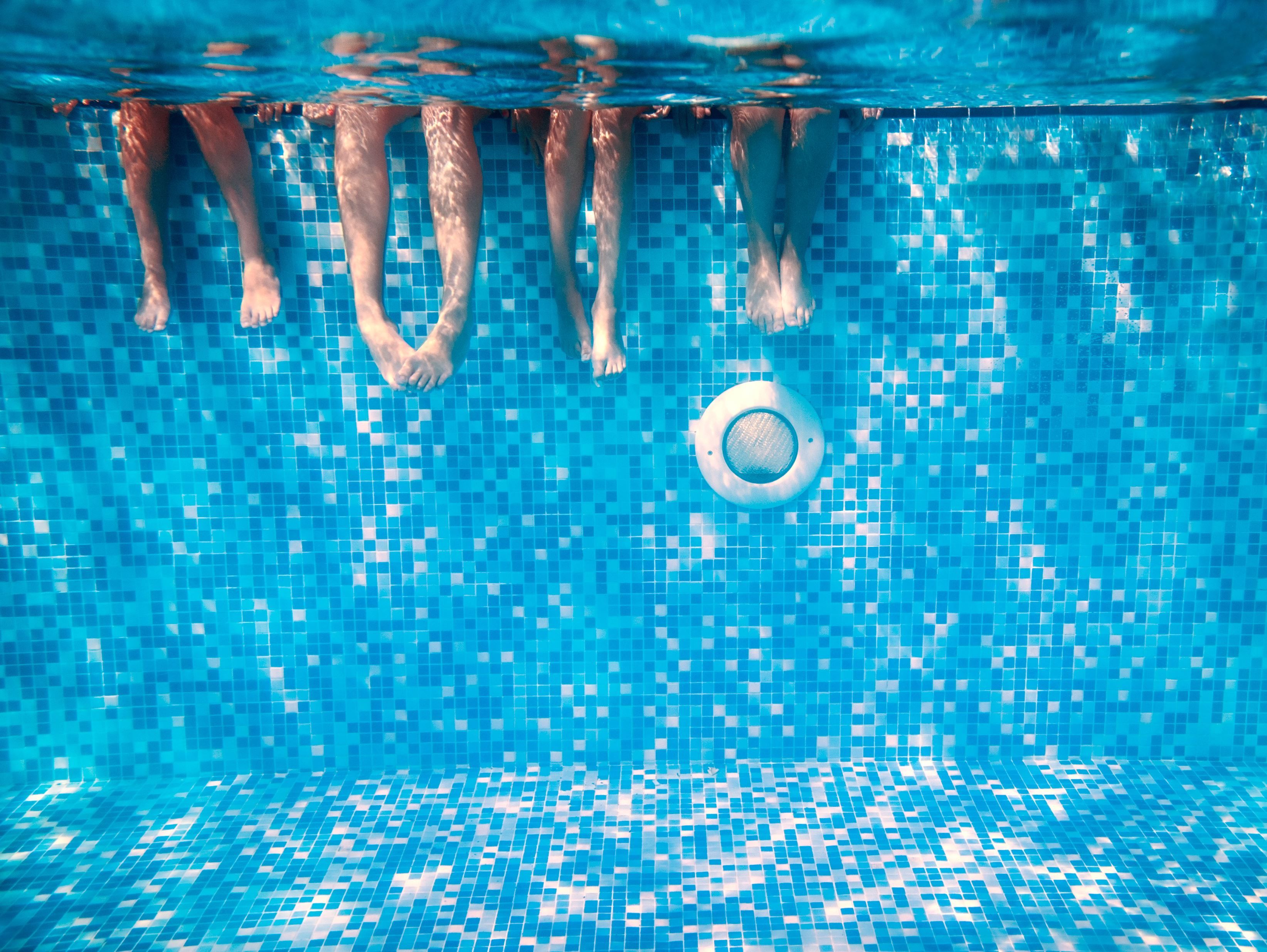 Underwater view of four people's legs in a pool with blue mosaic tiles