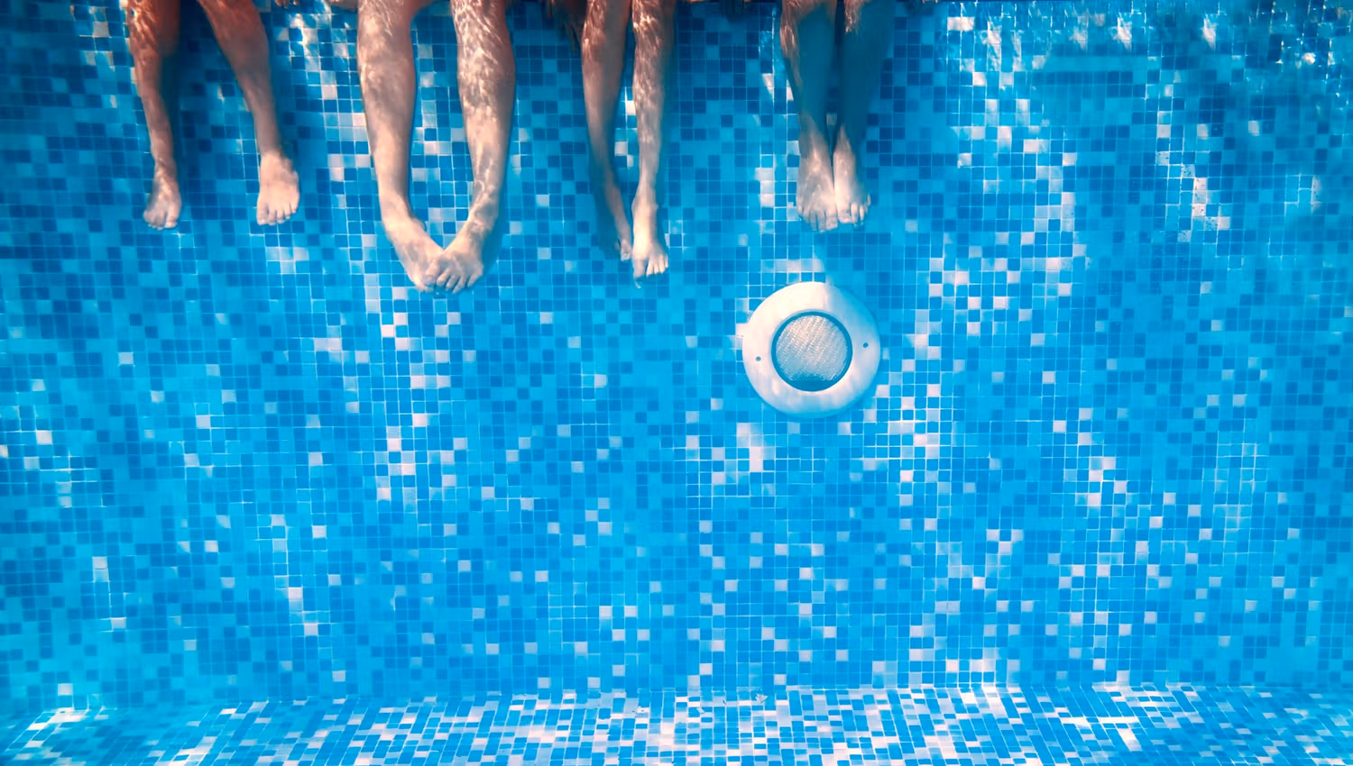 Underwater view of four people's legs in a pool with blue mosaic tiles