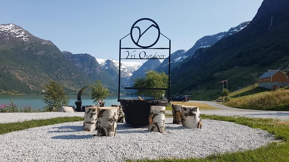 Gravel seating area with log stools and a metal fire pit under a sign, set by a lake with mountains behind.