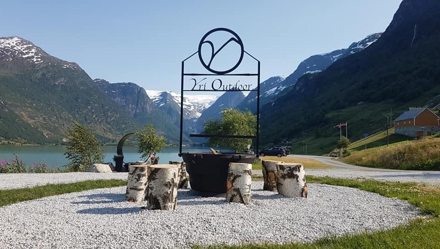Gravel seating area with log stools and a metal fire pit under a sign, set by a lake with mountains behind.