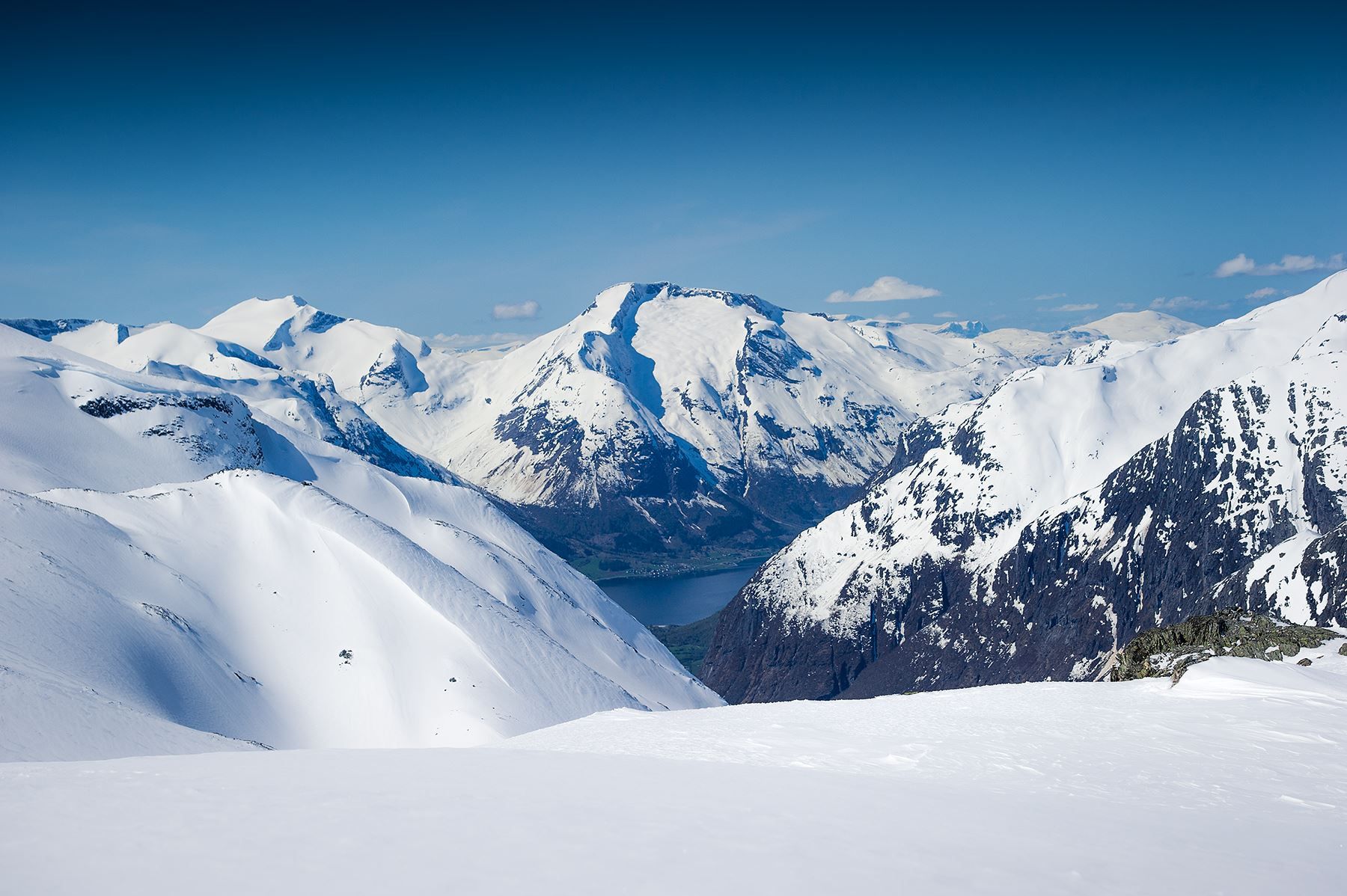 Wide view of snow-covered mountain peaks and ridges with dark rocky slopes and a lake in a distant valley.
