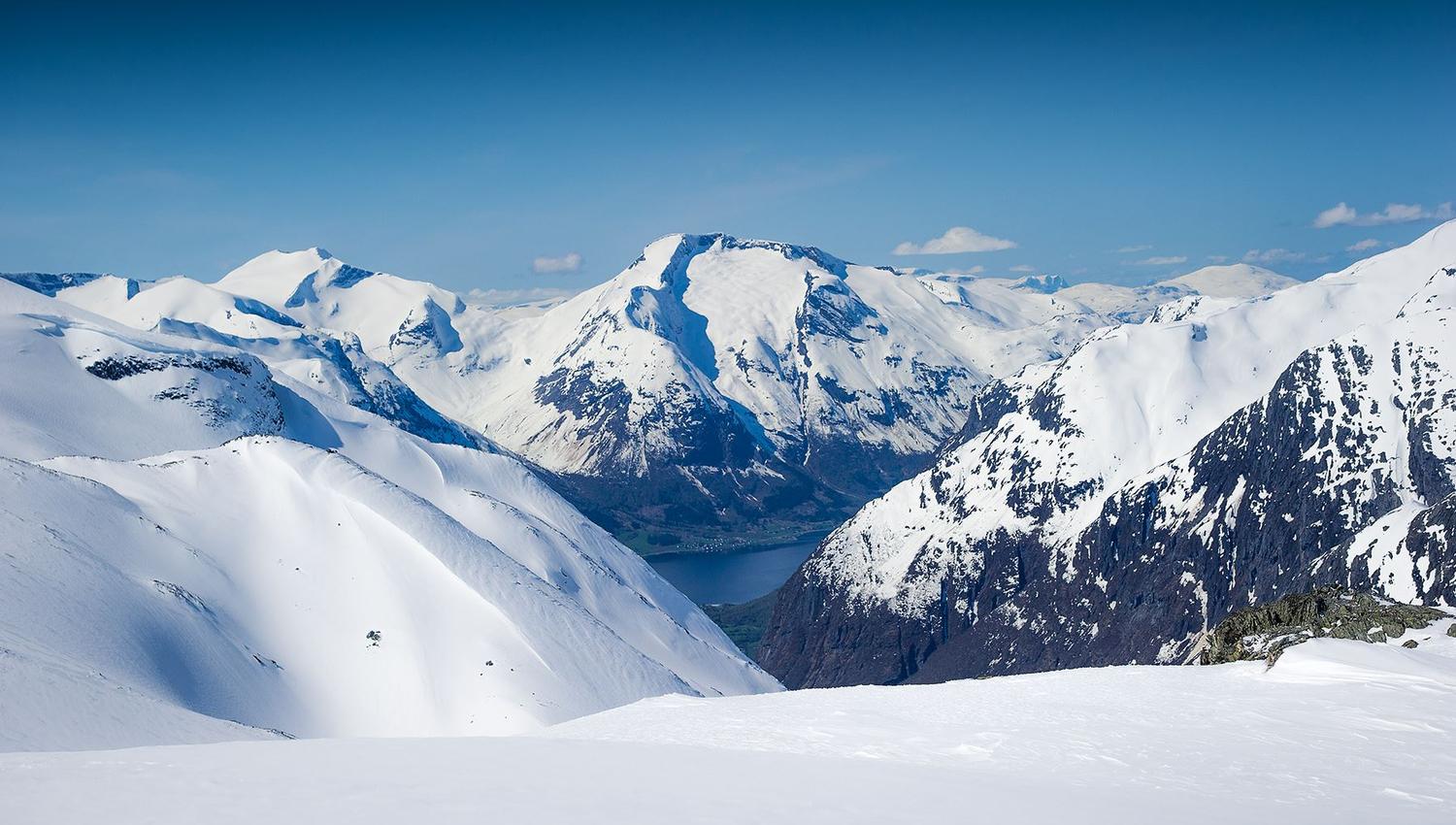 Wide view of snow-covered mountain peaks and ridges with dark rocky slopes and a lake in a distant valley.