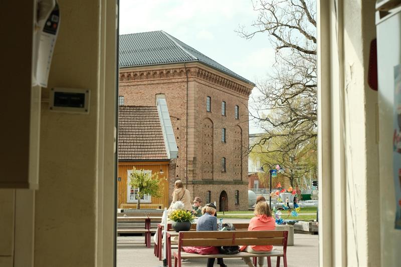 Gjester sitter ved utendørs kafébord på Karljohansvern, med en historisk mursteinsbygning i bakgrunnen.