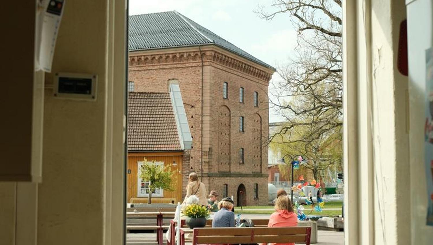Gjester sitter ved utendørs kafébord på Karljohansvern, med en historisk mursteinsbygning i bakgrunnen.