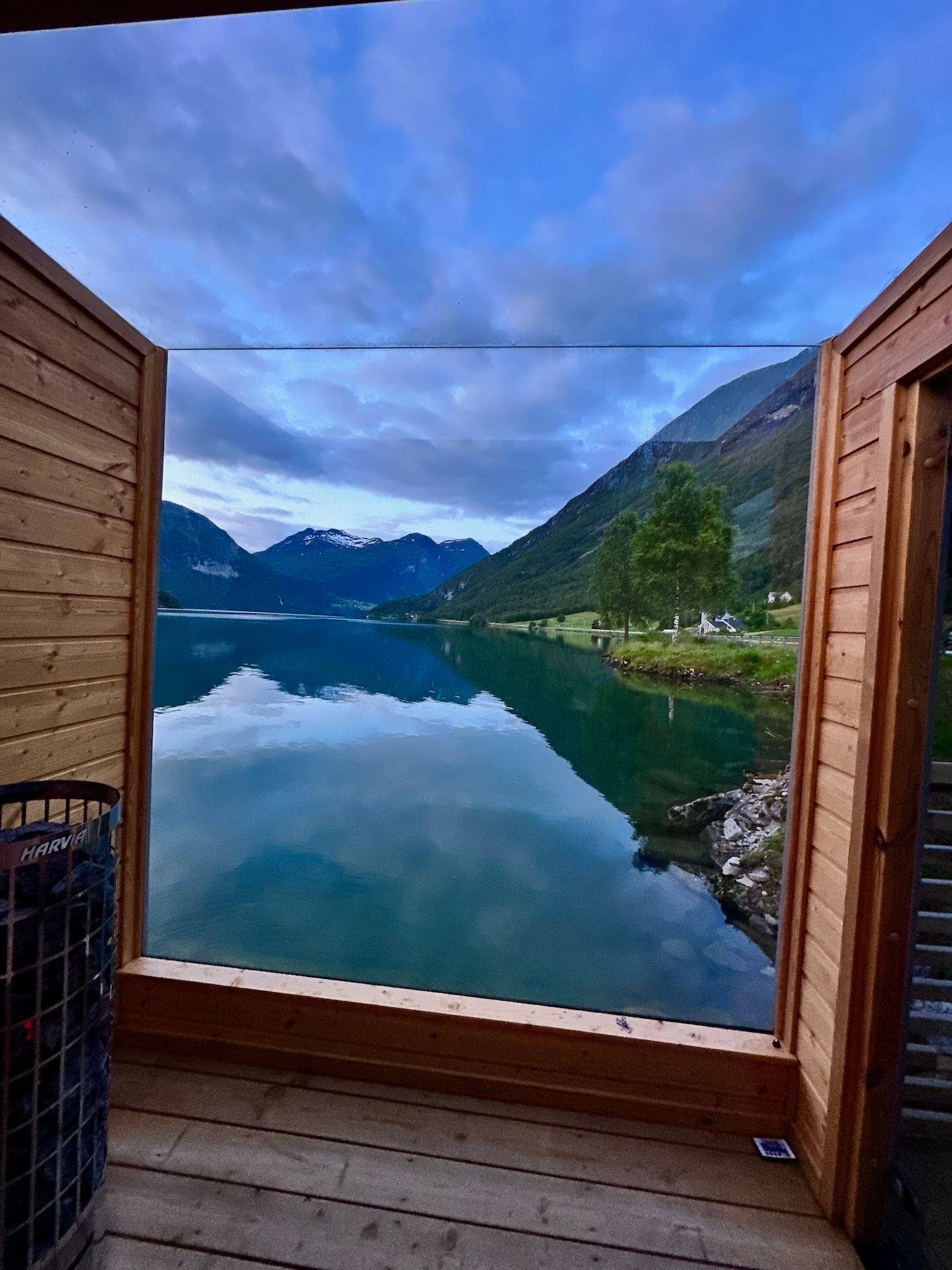 Wooden deck frames a calm mountain lake with cloud reflections, green slopes, a lone tree, and distant peaks..