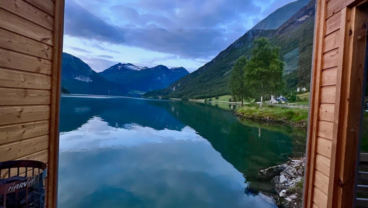 Wooden deck frames a calm mountain lake with cloud reflections, green slopes, a lone tree, and distant peaks..