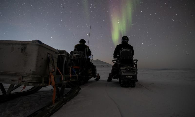 To personer sittende på snøscooter ser på nordlyset