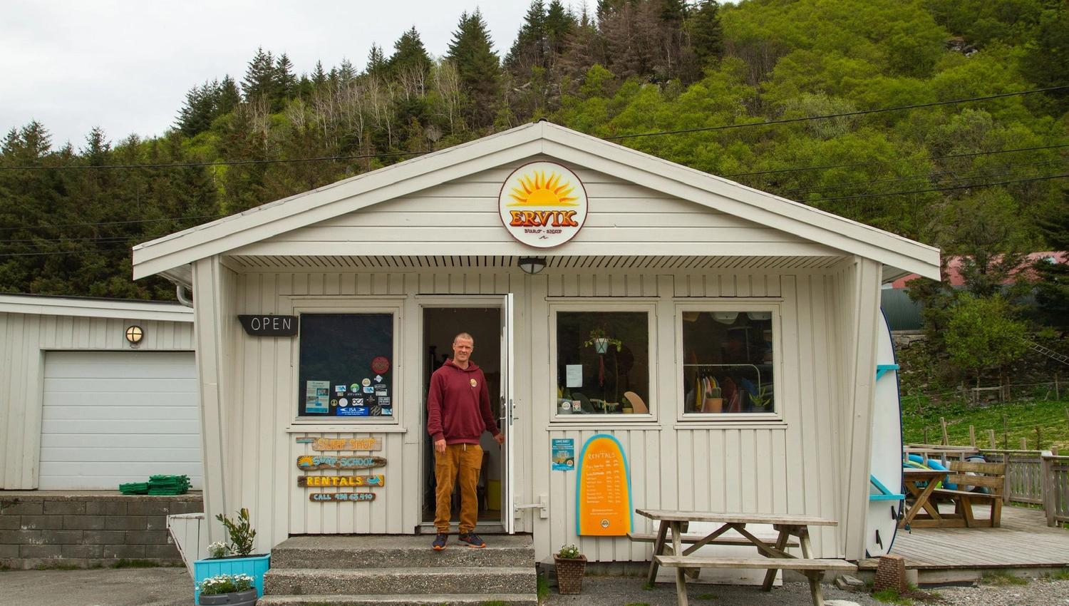Ein mann står i døropninga til ei lita surfebutikk, Ervik Surfshop, i ei kvit trebygning, med grøne åsar i bakgrunnen..