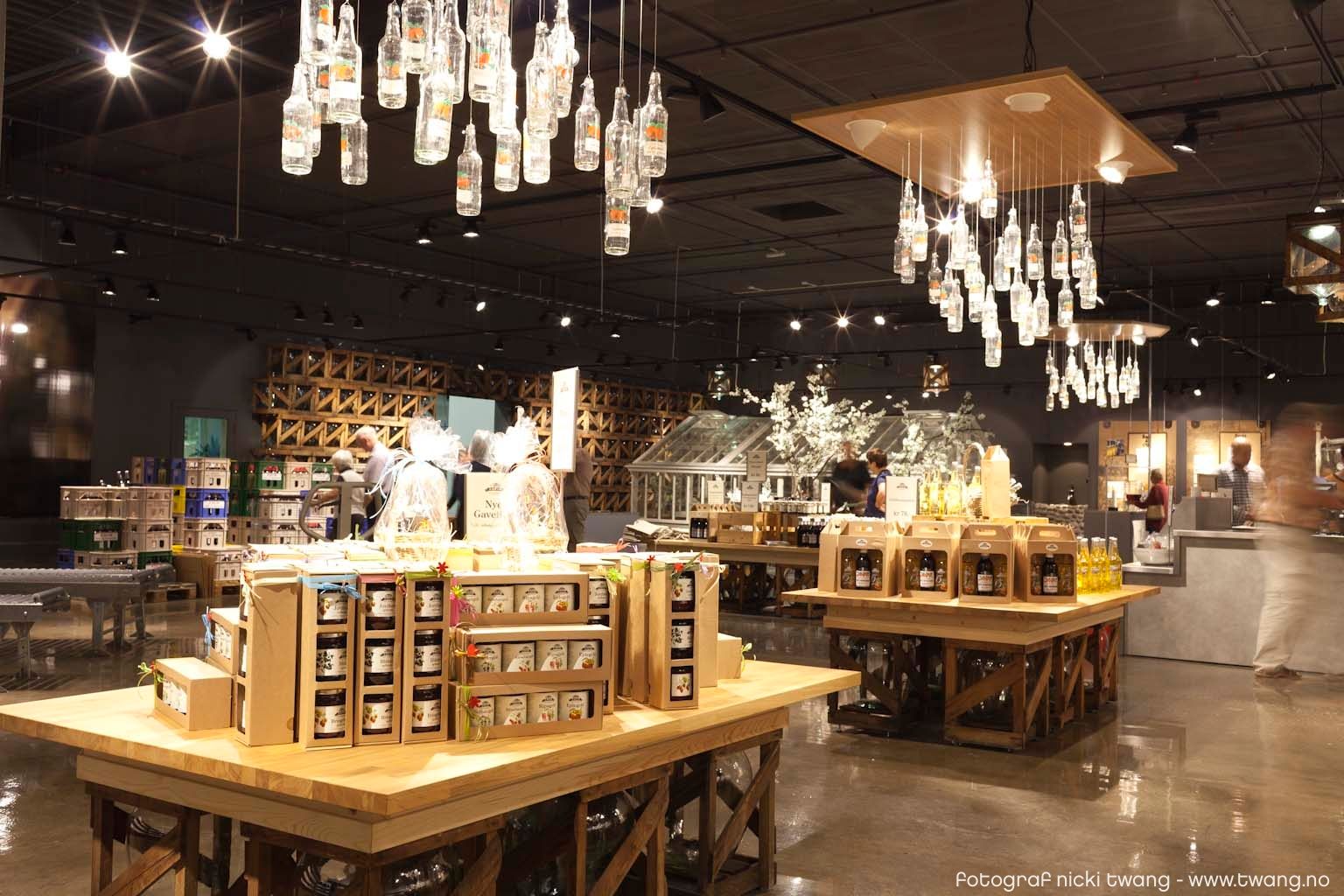 A modern shop interior with large wooden tables displaying local products such as jams, juices, and bottled drinks. Above the tables hang lights made 