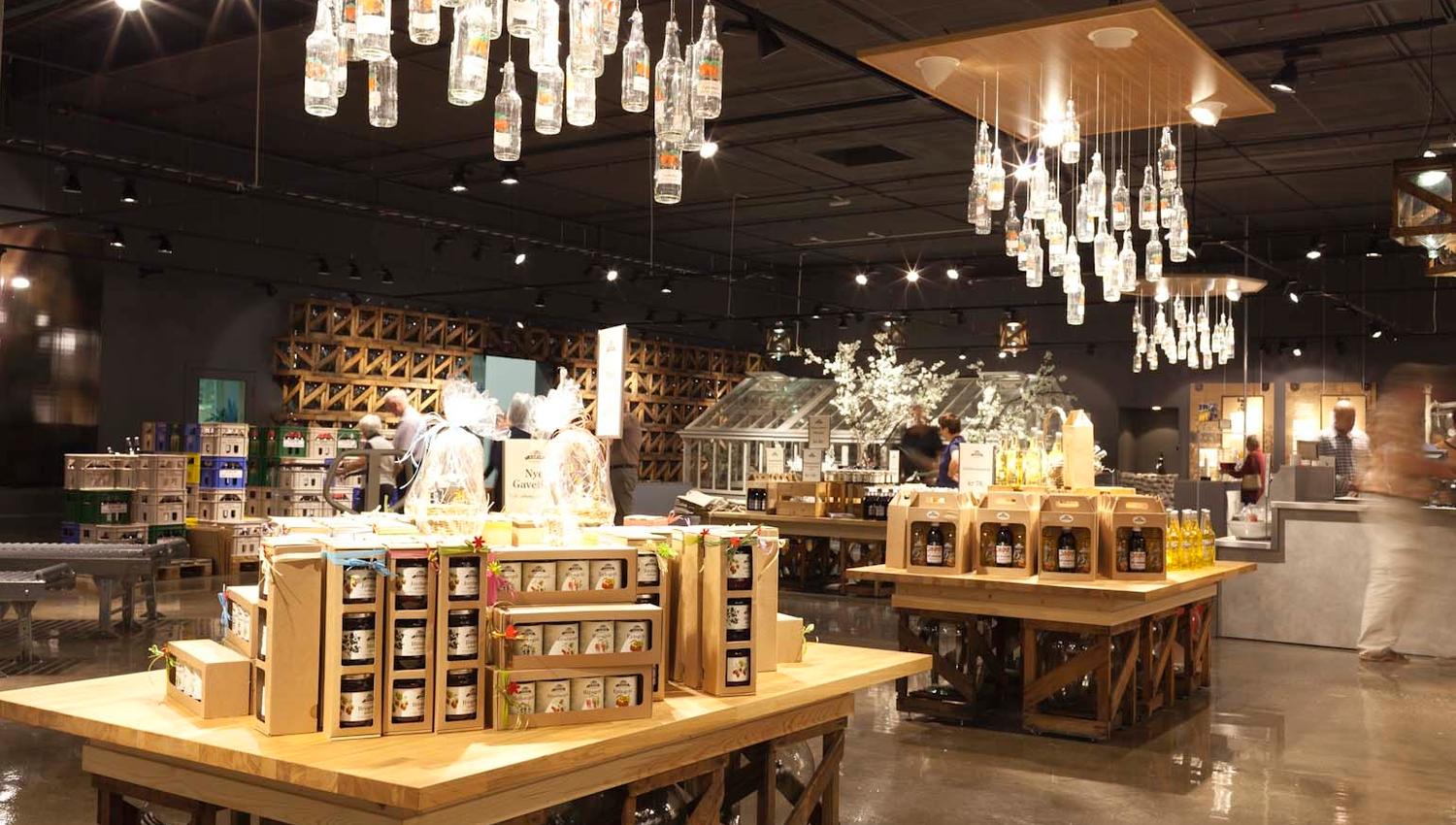 A modern shop interior with large wooden tables displaying local products such as jams, juices, and bottled drinks. Above the tables hang lights made