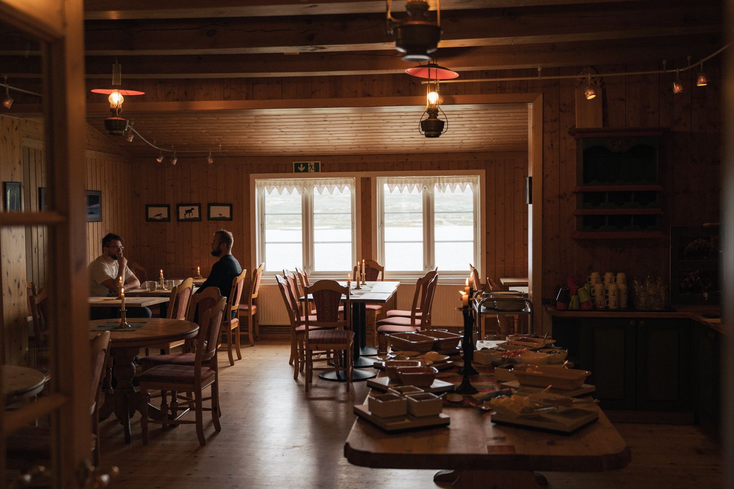 The dining room in dim light. Two people are sitting by the window and eating. The breakfast buffet is shown at the front.