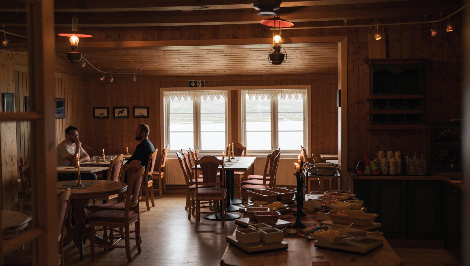 The dining room in dim light. Two people are sitting by the window and eating. The breakfast buffet is shown at the front.