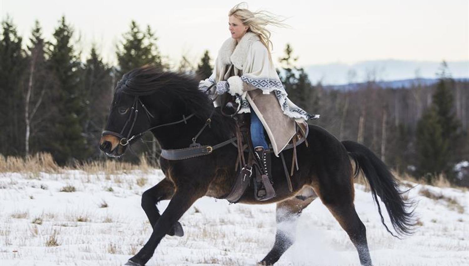 Kvinne rir på en svart hest over et snødekt jorde, med skog og fjell i bakgrunnen