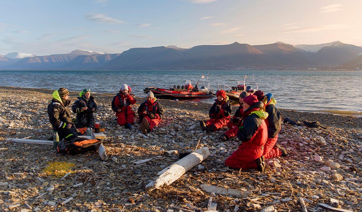 En gruppe med gjester og to guider som sitter i ring på en strand og prater sammen, med to RIB båter ankret langs sjøkanten og en fjord i bakgrunnen