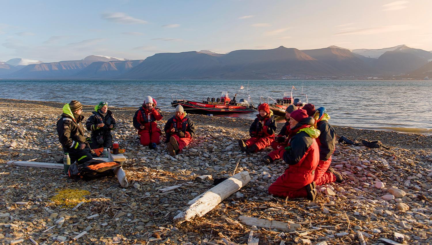 En gruppe med gjester og to guider som sitter i ring på en strand og prater sammen, med to RIB båter ankret langs sjøkanten og en fjord i bakgrunnen