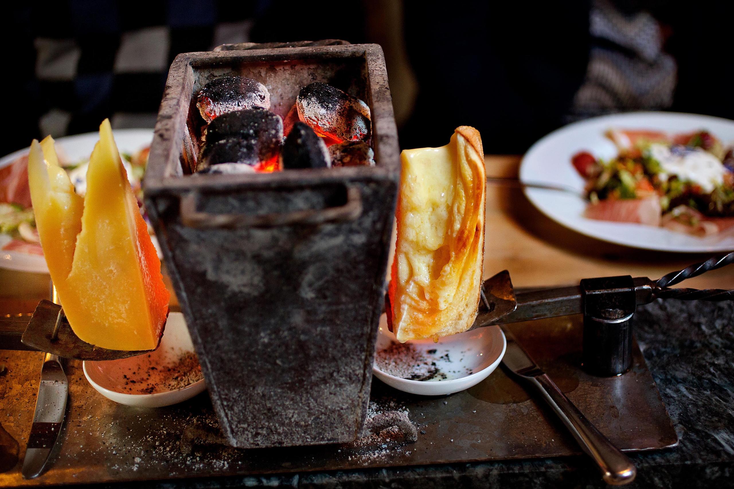 Tabletop raclette grill with glowing coals and melting cheese wedges on metal holders, plates of food behind..